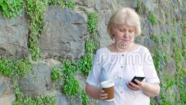 使用智能手机和喝咖啡的老年妇女。 背景中野生石头的复<strong>古墙</strong>。